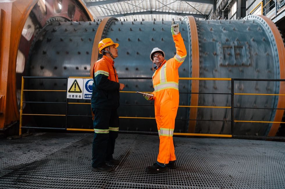 Two men in front of a mill at a mining site and the other pointing up. 